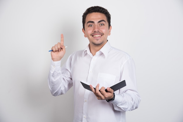 Positive businessman with notebook and pen pointing up on white background.