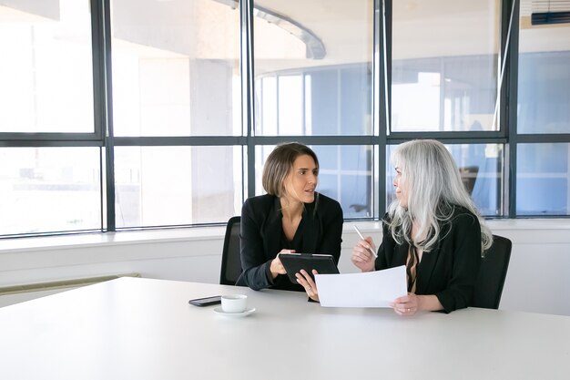 Positive businesswomen discussing and analyzing reports. Two female employees sitting together, holding documents, using tablet and talking. Teamwork concept