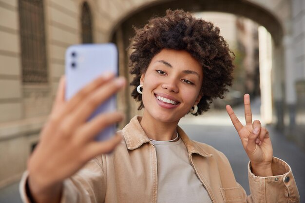 Positive carefree curly haired woman enjoys video call in city uses internet in roaming makes peace gesture smiles gladfully at camera wears stylish clothes poses against building background.