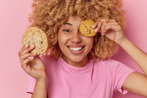Positive carefree curly haired woman has sweet tooth holds delicious cookies over eye smiles pleasantly dressed in casual t shirt glad to eat delicious snack isolated over pink studio background