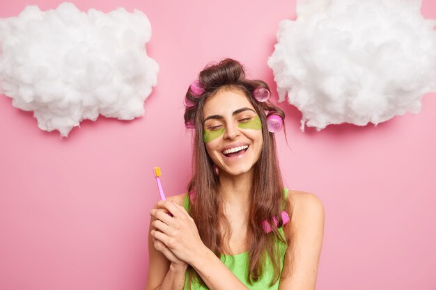 Positive carefree girl makes perfect hairstyle holds toothbrush going to clean teeth enjoys morning routines cares abot skin uses collagen pads smiles toothily isolated over pink wall clouds up