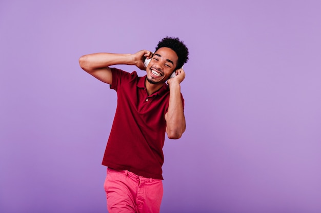 Positive carefree man laughing while enjoying song. Well-dressed guy smiling and touching his headphones.