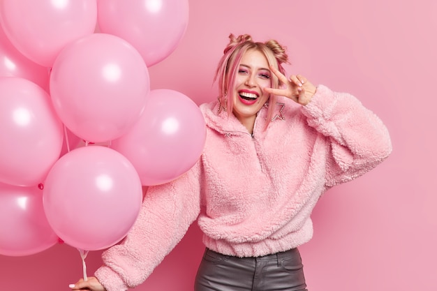 Positive carefree stylish millennial girl dressed in warm coat makes peace sign over eye smiles gladfully has party mood enjoys holiday holds bunch of inflated balloons