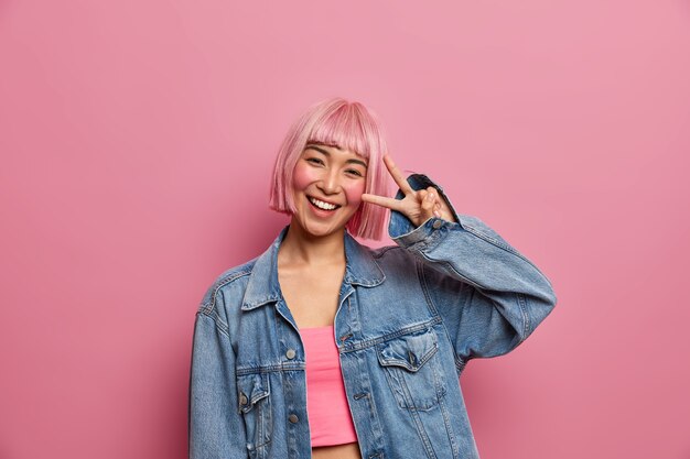 Positive carefree teenage girl with pink hairstyle shows victory peace gesture or disco sign, enjoys good day and smiles with teeth, wears fashionable clothes, feels happy, 