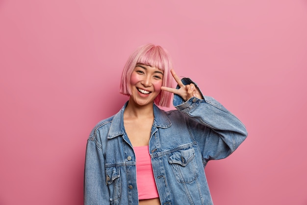 Positive carefree teenage girl with pink hairstyle shows victory peace gesture or disco sign, enjoys good day and smiles with teeth, wears fashionable clothes, feels happy, 