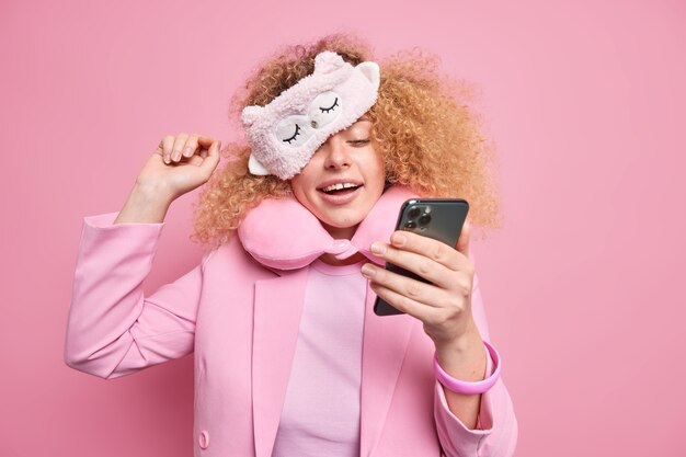 Positive carefree woman addicted to modern technologies checks newsfeed in social networks via smartphone after awakening wears soft sleepmask formal clothes chats online isolated on pink wall