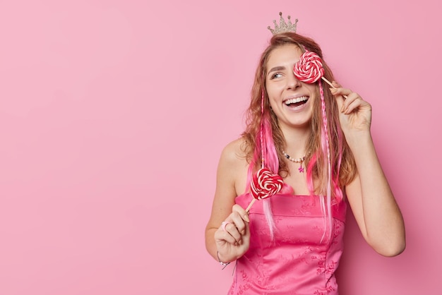 Positive carefree woman covers eye with heart shaped lollipop smiles gladfully wears small crown and dress looks away isolated over pink background with copy space for your promotional content