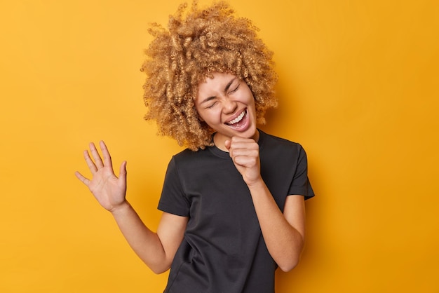Positive carefree woman dances and sings song holds hand near mouth as if microphone wears casual black t shirt isolated over vivid yellow background has upbeat mood People and emotions concept