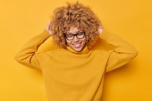 Free photo positive carefree woman has fun keeps hands on head smiles gently closes eyes from satisfaction wears spectacles and long sleeved jumper recalls something nice isolated over yellow background