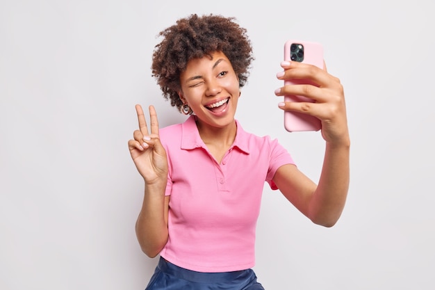 Positive carefree woman has online conversation via smartphone makes peace gesture winks eye smiles broadly makes Portrait of herself dressed casually isolated over white wall