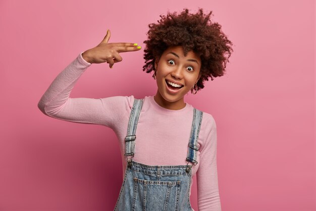 Positive carefree woman shoots with finger gun gesture at temple, commits suicide, foolishes around, looks joyfully , wears denim overalls and poloneck, isolated on pink pastel wall