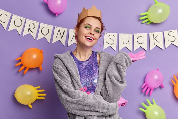 Positive carefree woman with bright makeup wears crown dressing gown and rubber gloves being in good mood during domestic party poses against garland and inflated balloons