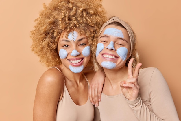 Positive carefree young women with beauty nourishing mask on face stand closely to each other smile broadly make peace gesture undergo skin care treatments isolated over brown studio background