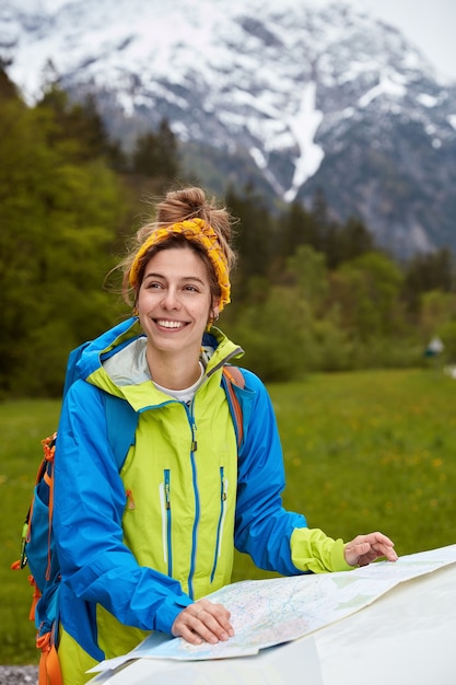 Позитивная кавказская женщина-путешественница стоит возле карты, изучает маршрут, носит желтый шарф на голове, повседневный анорак.