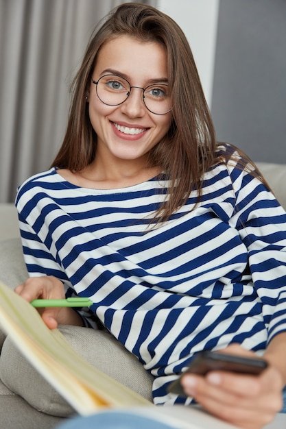 Positive Caucasian girl does paper work, writes notes, uses mobile phone