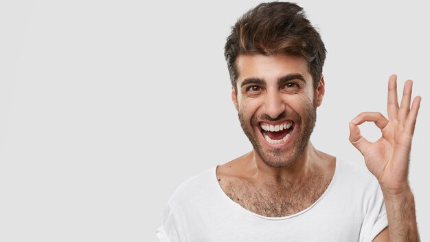 Positive Caucasian male shows okay or zero sign, smiles with excitement, feels happy