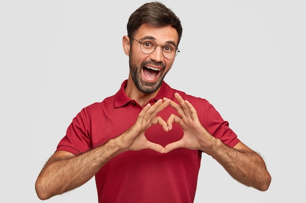 Positive Caucasian male with glad expression, shows heart gesture over chest, expresses friendly attitude and love, wears red bright t-shirt, isolated over white wall