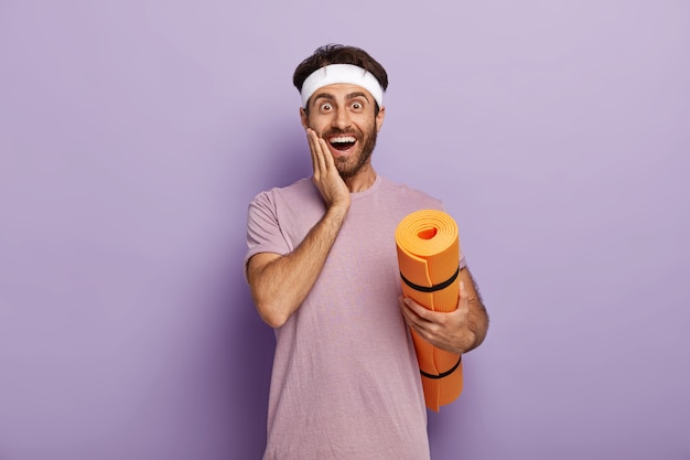 Positive Caucasian man stands with rolled up mat, touches cheek, wears headband and t shirt, stands against purple wall has fitness workout regularly, waits for coach, motivated for training