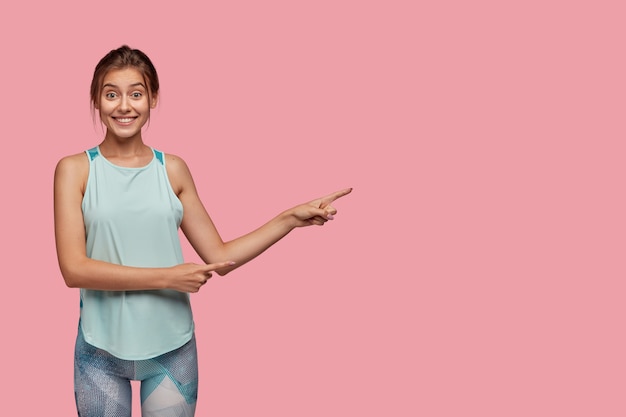 Positive Caucasian woman with cheerful expression, dressed in casual vest and leggings, points aside with both index fingers, stands against pink wall, has healthy lifestyle, likes sport