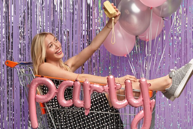 Positive Caucasian woman with light hair, poses at trolley, makes selfie with little camera, wears polka dot dress and sneakers, has fun at party