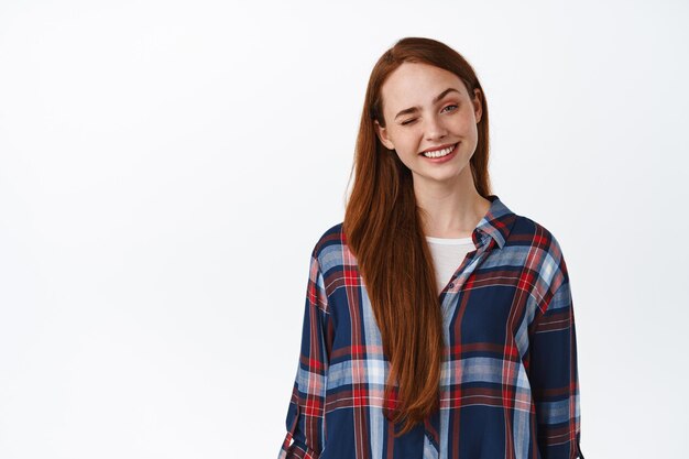 Positive caucasian woman with red long hair, freckles and clean skin, smiling and winking at camera happy, standing in plaid shirt against white background