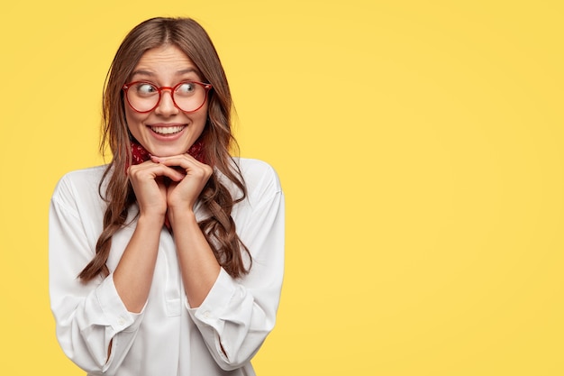 Positive Caucasian woman with tender smile, keeps hands under chin, giggles positively aside, dressed in white shirt, stands against yellow wall with free space for your promotional content