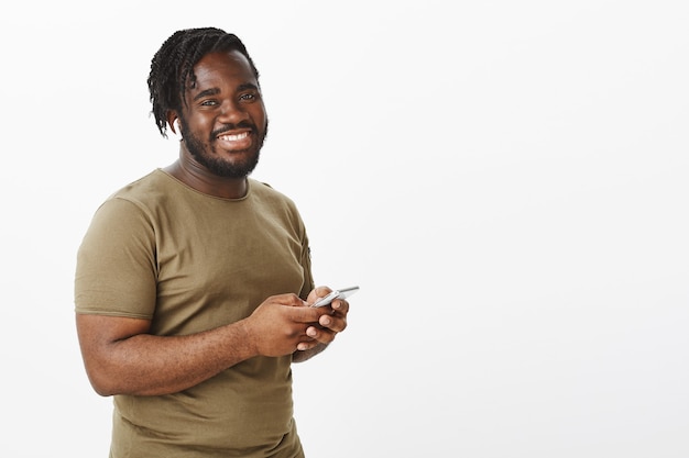 Positive charming guy in a brown t-shirt posing against the white wall with his phone