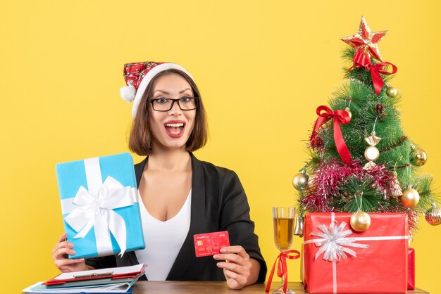 Positive charming lady in suit with santa claus hat and eyeglasses showing gift and bank card in the office on yellow isolated 