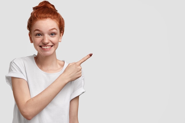 Positive charming red haired woman shows her house, points with index finger aside, being glad and suggests use copy space, dressed casually, models