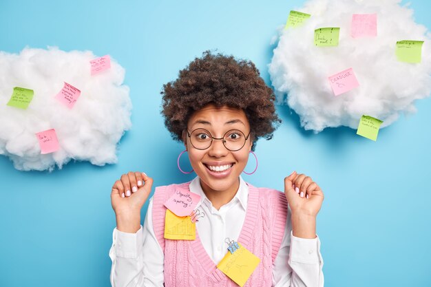 Positive cheerful Afro American woman raises hand looks excitedly at front glad to hear praise for good work wears round spectacles formal clothes surrounded by sticky notes with written tasks
