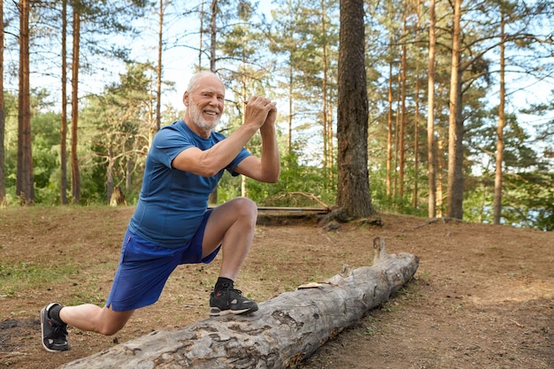 Positive cheerful European male pensioner in t-shirt, shorts and sneakers having warm up routine outdoors, standing on log with one feet, holding hands in front of him, doing lunges, smiling