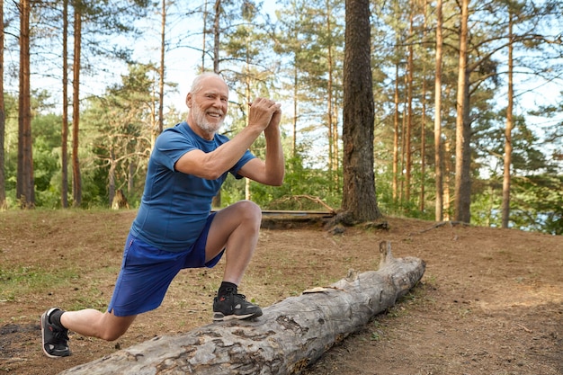 Позитивный жизнерадостный европейский пенсионер в футболке, шортах и кроссовках, разогревающий рутину на открытом воздухе, стоит на бревне одной ногой, держась за руки перед собой, делает выпады, улыбается