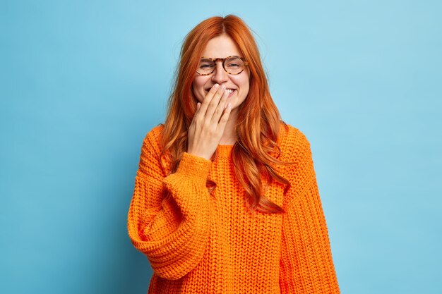 Positive cheerful redhead woman smiles happily tries to hide emotions covers mouth with hand feels shy hears hilarious joke wears knitted sweater.