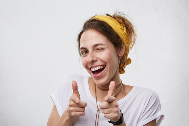 Positive cheerful young woman wearing yellow scarf on head and white casual T-shirt blinking her eyes and smiling pointing with index fingers. Happy attractive woman pointing at you