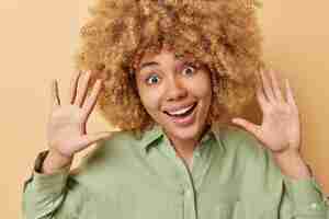 Free photo positive cheerful young woman with blonde curly bushy hair has glad expression keeps palms raised towards camera dressed in casual shirt isolated over brown background human emotions concept