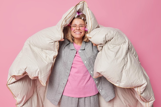 Positive cheerful young woman wrapped in duvet has happy expression after healthy sleep wakes up in good mood wears comfortable pajama makes hairstyle with rollers poses against pink background