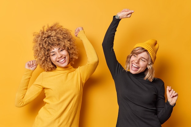Free photo positive cheerful young women have fun and dance carefree shake arms dressed in casual turtlenecks have glad expressions isolated over yellow background people happiness and emotions concept
