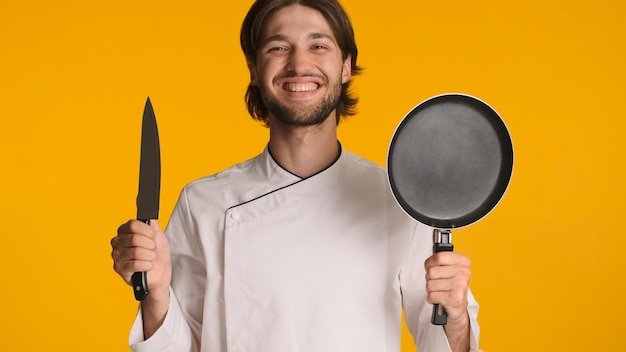 Positive chef dressed in uniform holding knife and frying pan looking cheerful at camera over yellow background Young man with cook equipment in hands ready to work