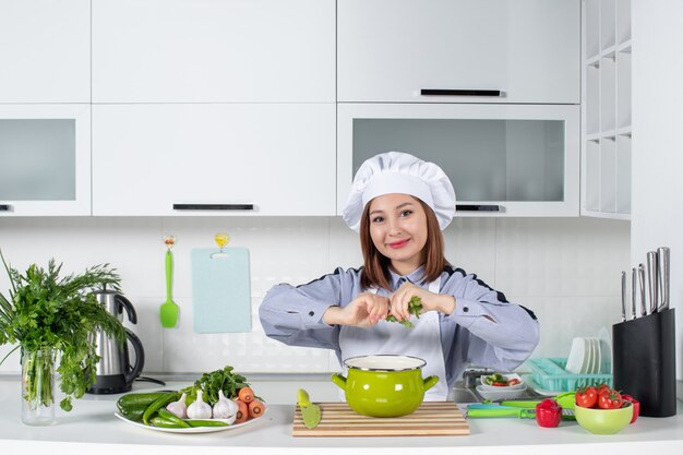 Позитивный повар и свежие овощи с кухонным оборудованием и добавление зелени в кастрюлю на белой кухне