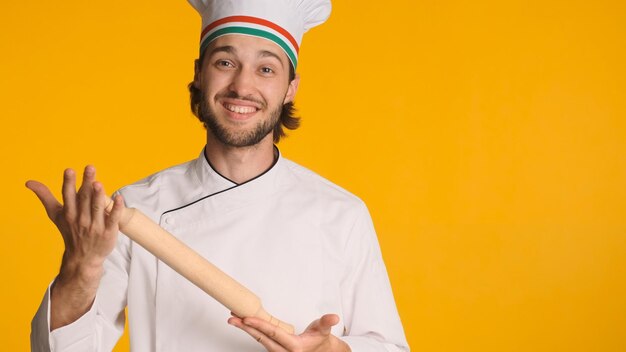 Positive chef in white uniform and cap with wooden rolling pin in hands posing at camera over colorful background