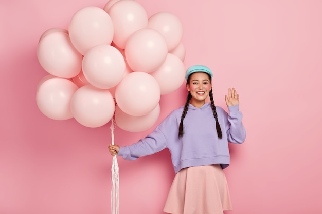 Positive Chinese woman comes on friends birthday party, greets fellows, has dark hair combed in two plaits, dressed in casual outfit