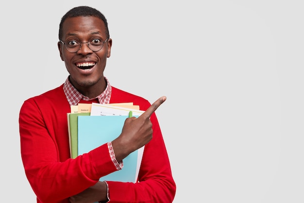 Free photo positive college student has dark skin, carries folders and book, points with cheerful expression aside, has toothy smile