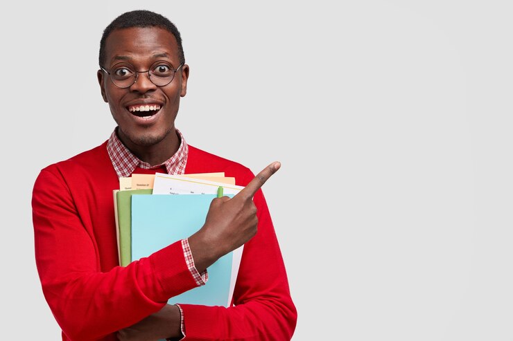 Positive college student has dark skin, carries folders and book, points with cheerful expression aside, has toothy smile