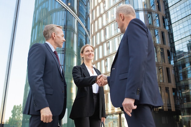 Positive confident business people meeting in city, shaking hands near office building. Low angle shot. Communication and partnership concept