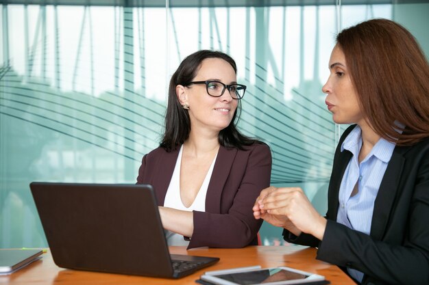 Positive confident young businesswomen discussing project while sitting at open laptop, talking and smiling.