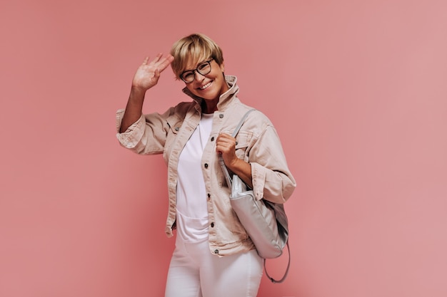 Позитивная крутая дама со светлой прической и современными очками в легкой одежде улыбается и машет рукой на розовом изолированном фоне.