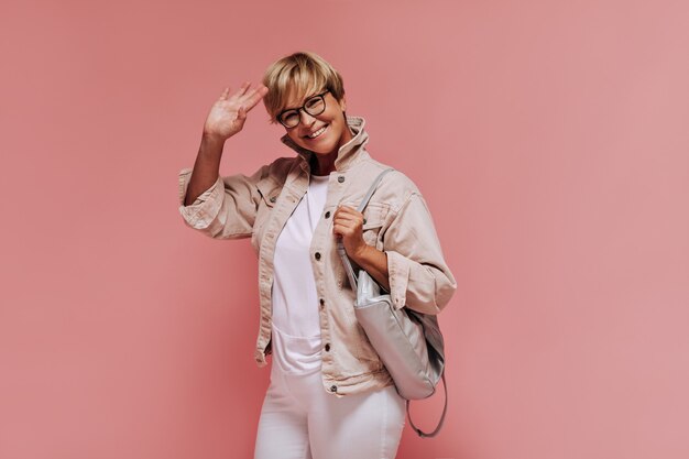 Positive cool lady with blonde hairstyle and modern glasses in light clothes smiling and waving hand on pink isolated backdrop. 