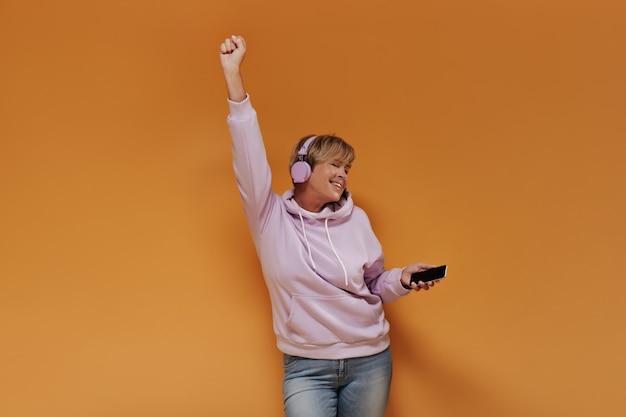 Positive cool lady with short hairstyle and lilac cool headphones in wide pink hoodie dancing and listening to music on isolated backdrop. 