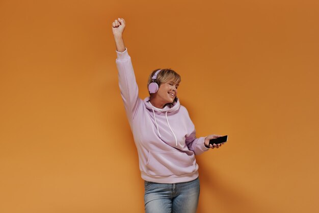 Positive cool lady with short hairstyle and lilac cool headphones in wide pink hoodie dancing and listening to music on isolated backdrop. 