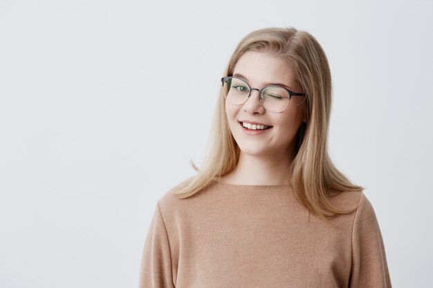 Positive coquettish European female student with blonde straight hair smiling happily, blinking  in a playful manner, flirting with you. Human facial expressions and emotions